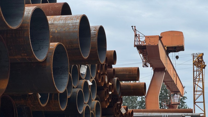 Tubos de acero apilados en un patio industrial, con una grúa puente de fondo, de cerca