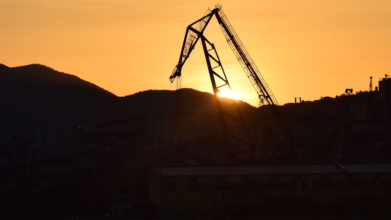 Silueta de una grúa portuaria contra un atardecer anaranjado, con montañas de fondo