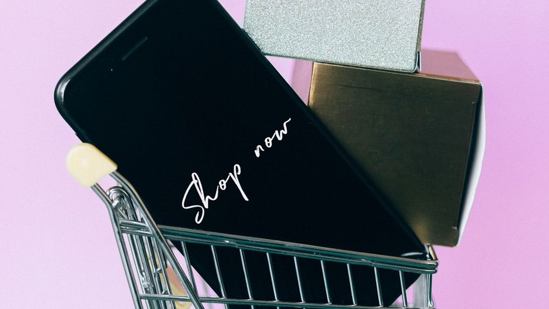 Celular con la pantalla de una app de compras dentro de un carrito de compras en miniatura, junto a cajas de regalo