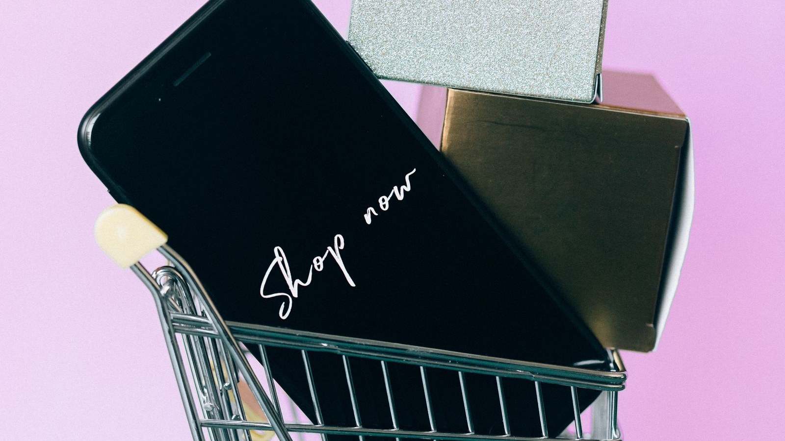 Celular con la pantalla de una app de compras dentro de un carrito de compras en miniatura, junto a cajas de regalo