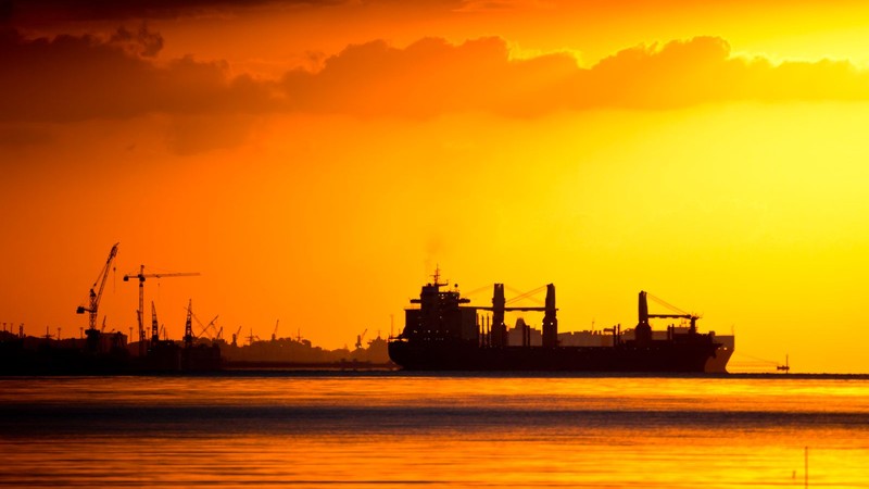 Silueta de un buque de carga navegando contra un atardecer intenso, con grúas portuarias de fondo
